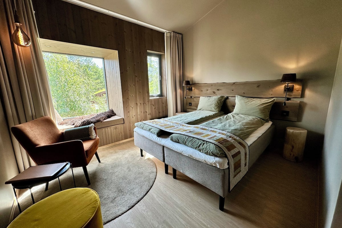 A bedroom with a bed, a chair, a small table, a lamp, and a window with curtains.
