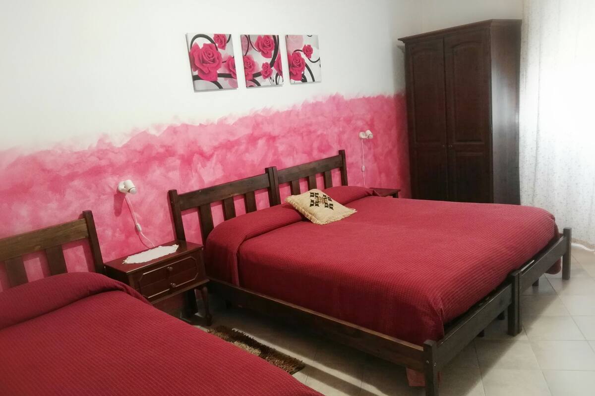 A bedroom with two wooden beds, each with a red bedspread and a decorative pillow. There is a nightstand with a lamp and a small table with a decorative item. The wall is painted pink, and there are three framed pictures above the beds.