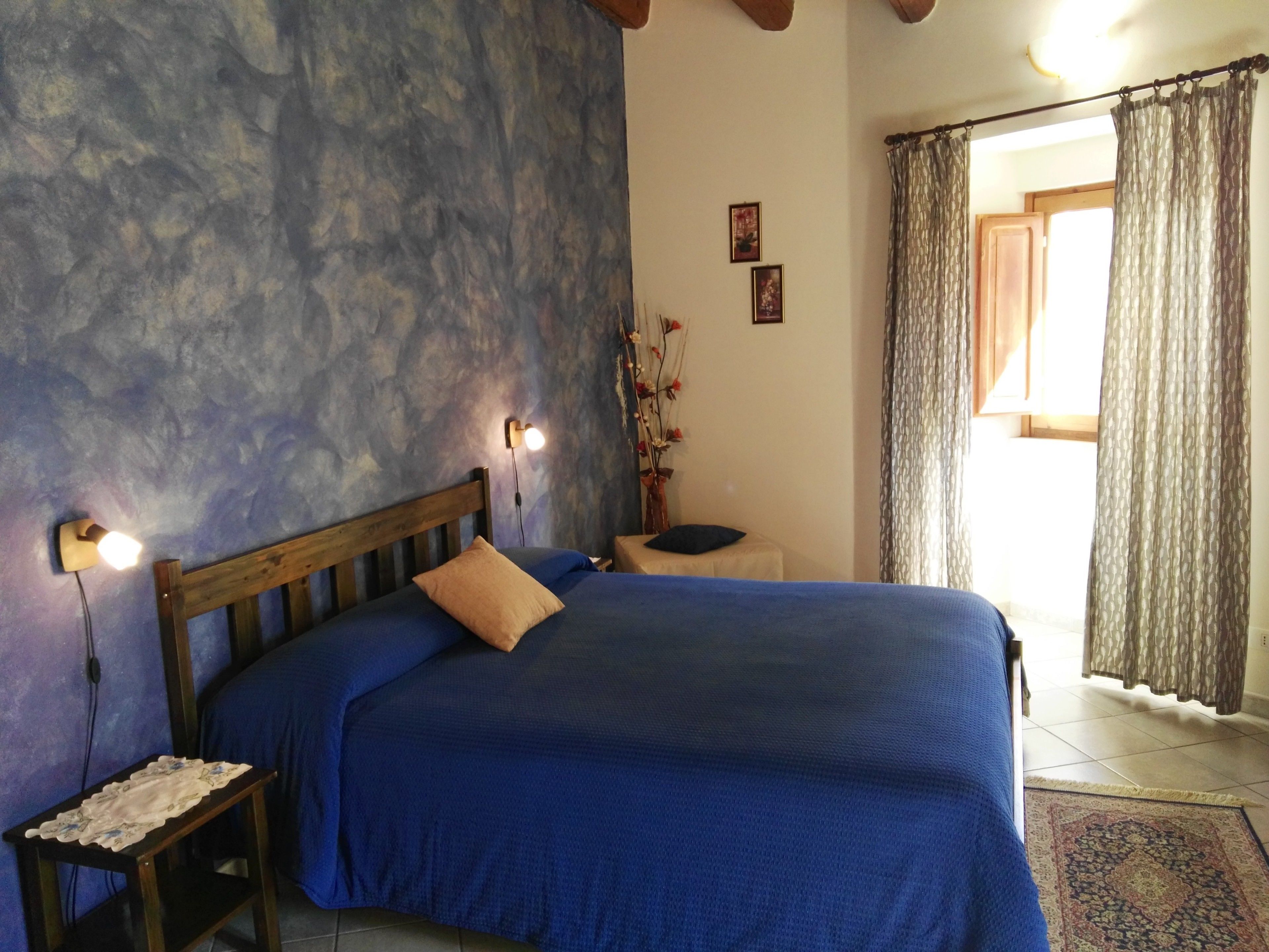A bedroom with a wooden headboard, a blue bedspread, a bedside table with a lamp, and a view of the outside through a window with curtains.