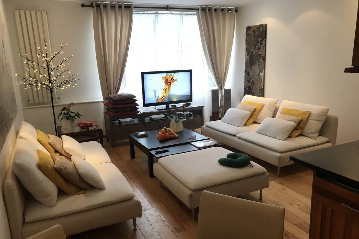 Una sala moderna con sofá, reposapiés y mesa de centro. Un televisor está montado en la pared y hay una planta en la mesita de noche.