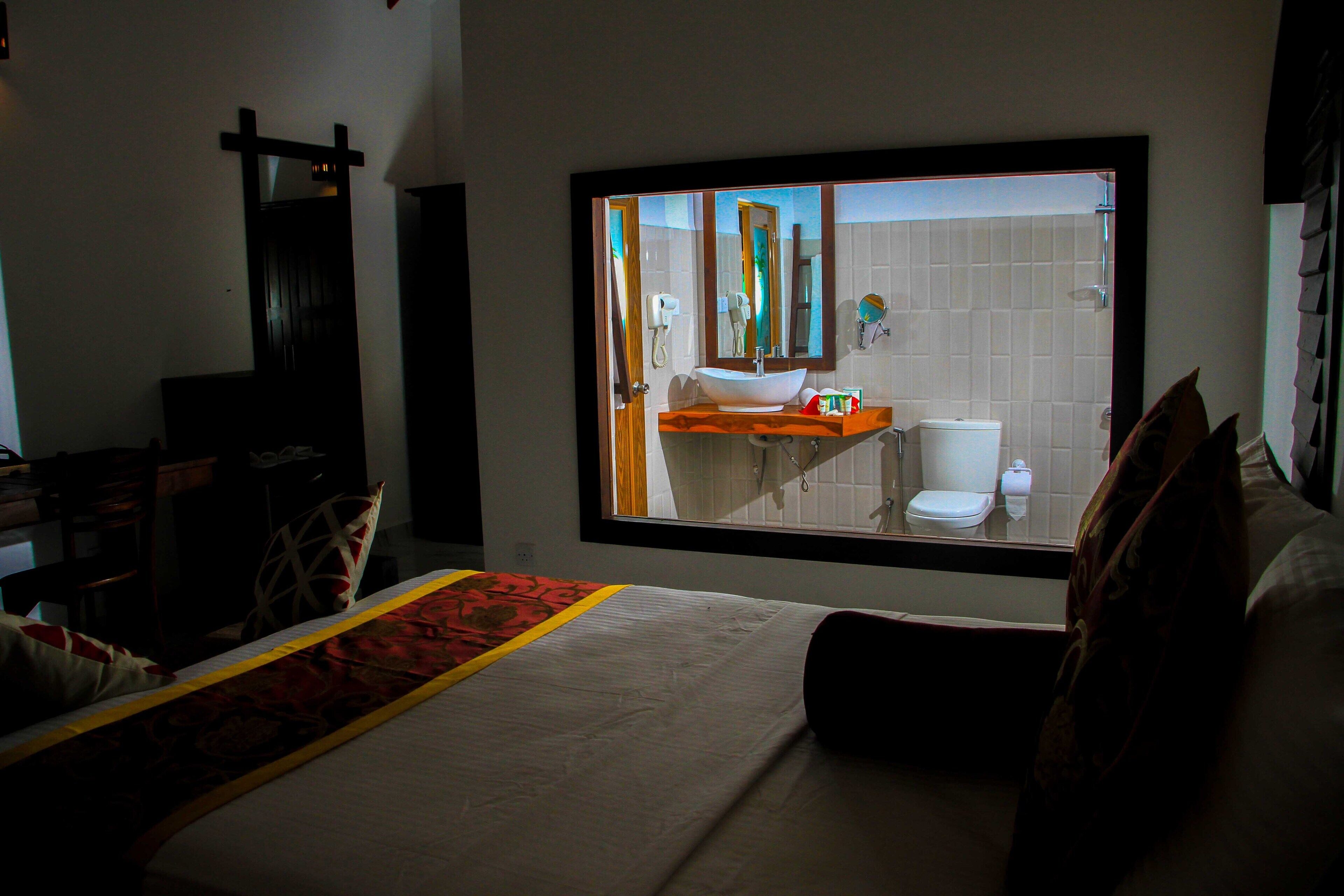 Un dormitorio con un ventanal grande, una cama con colcha roja y blanca, cabecera de madera y vistas a un baño con inodoro y lavamanos.