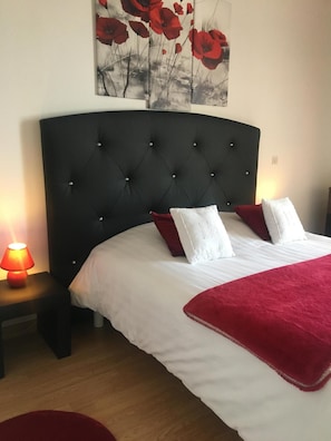 A bedroom with a tufted headboard, white bedding, red pillows, a red throw blanket, and a nightstand with a lamp.