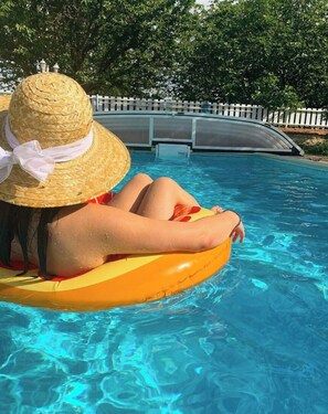 Eine Person entspannt sich auf einem gelben Schwimmding in einem klaren, blauen Pool, im Hintergrund ein weißer Zaun und Bäume.