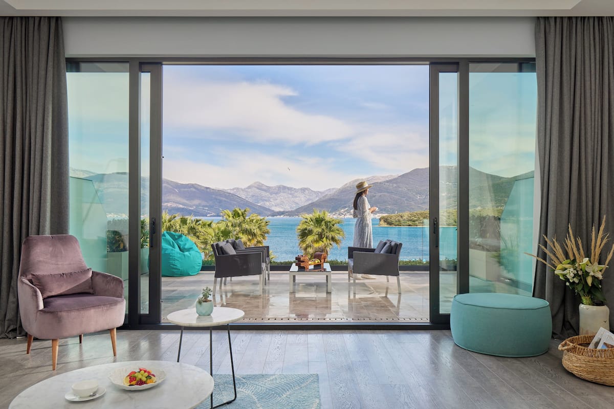 A modern living room with a large sliding glass door leading to a balcony with a scenic mountain and lake view.