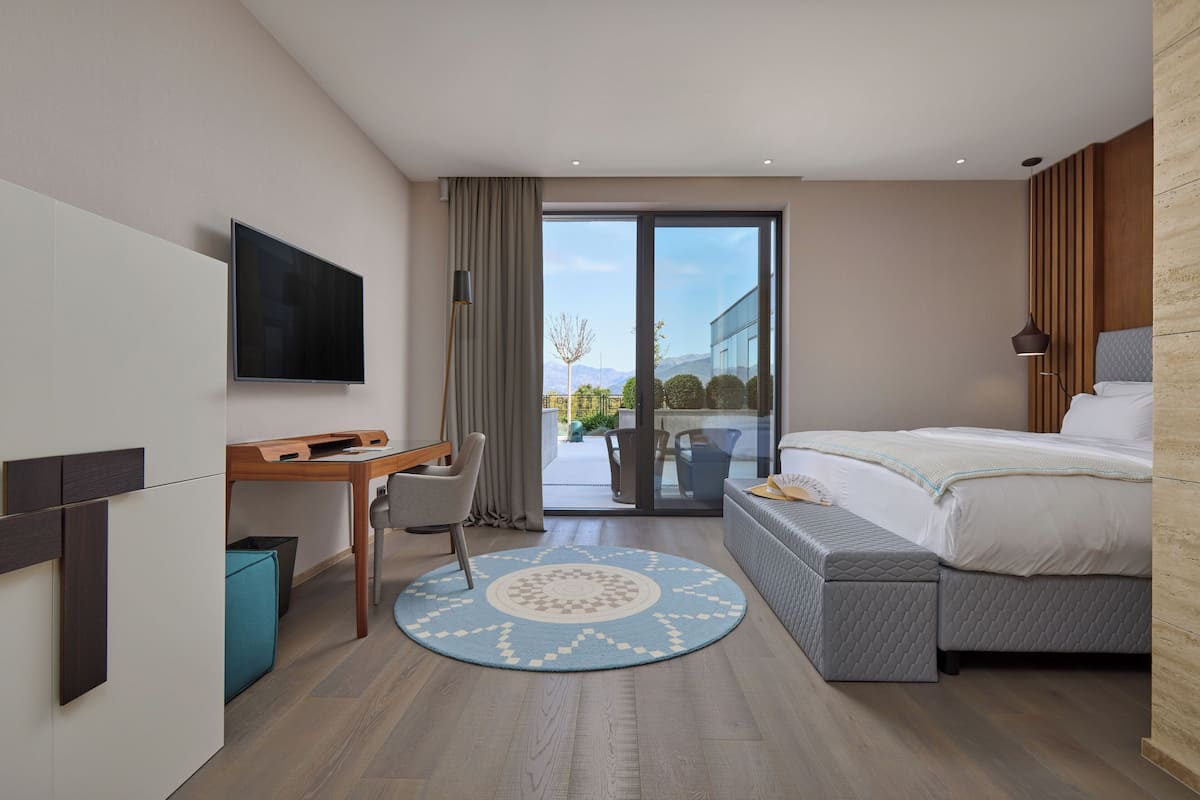 A modern hotel room with a bed, a desk, a chair, a TV, and a view of the outdoors.