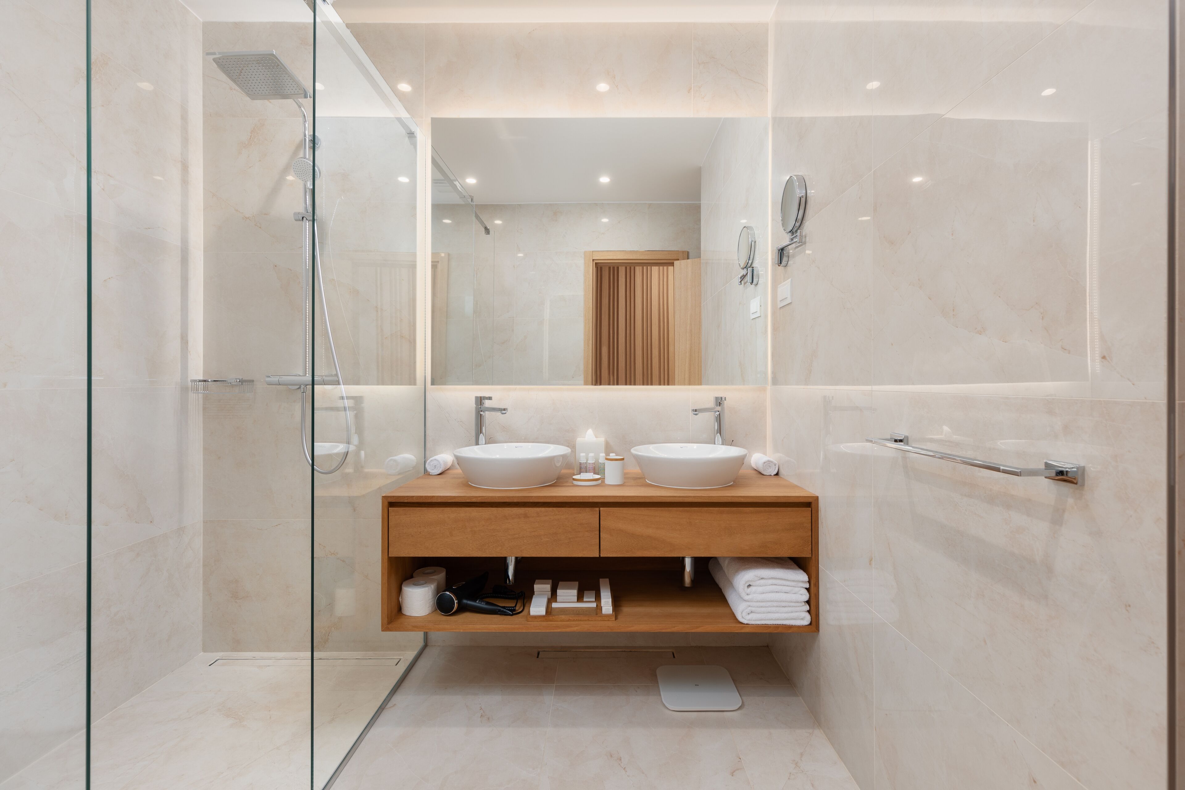 Ein modernes Badezimmer mit einer gläsernen Duschkabine, einem Holzbadezimmerhocker mit zwei Waschbecken und einem großen Spiegel.