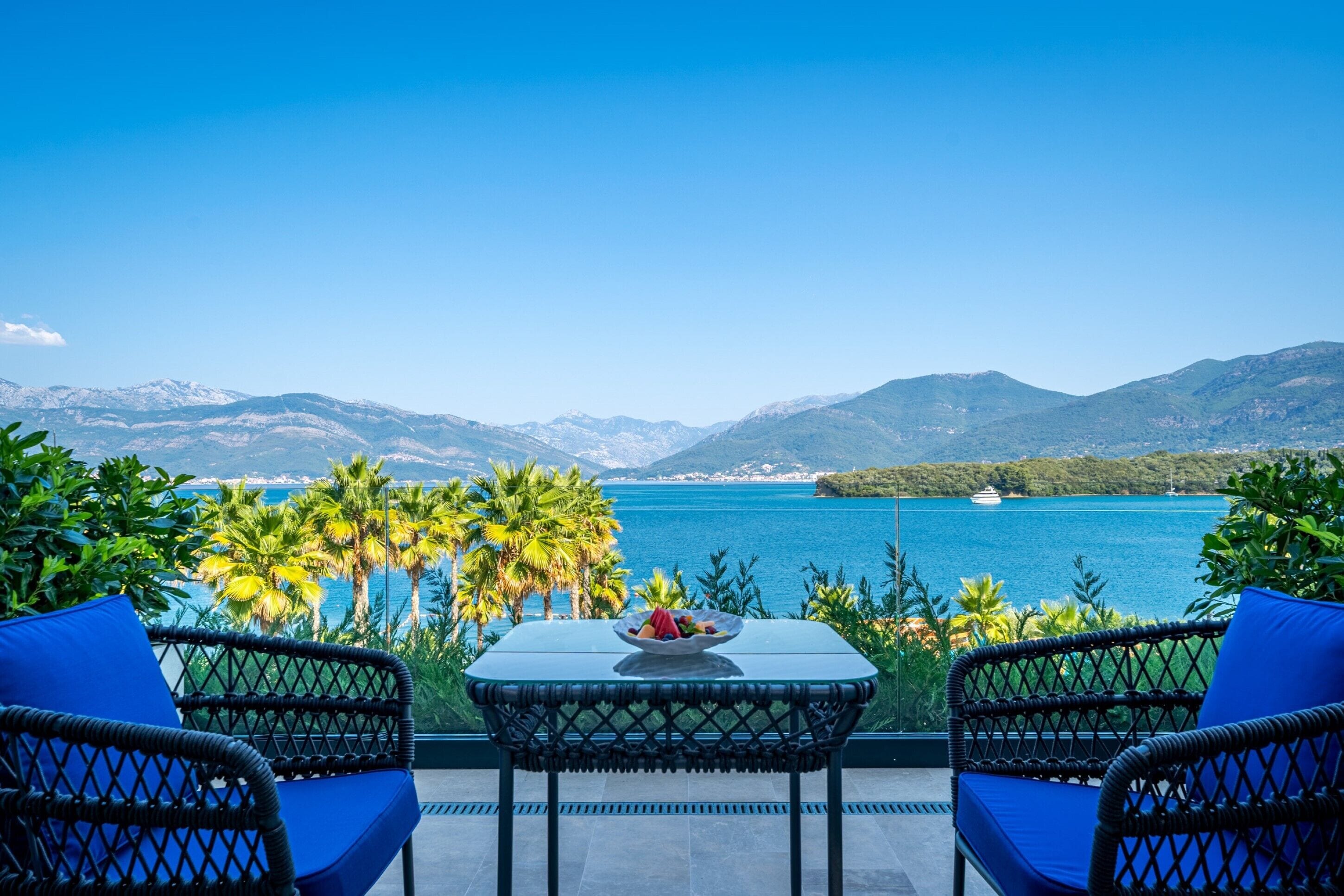 A terrace with wicker chairs and a table, overlooking a lake with mountains in the background.