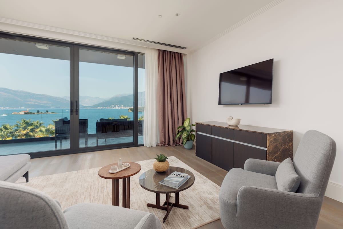 A modern living room with a large sliding glass door leading to a balcony with a sea view. There is a flat-screen TV mounted on the wall, a small round table with a plant, and a grey armchair.
