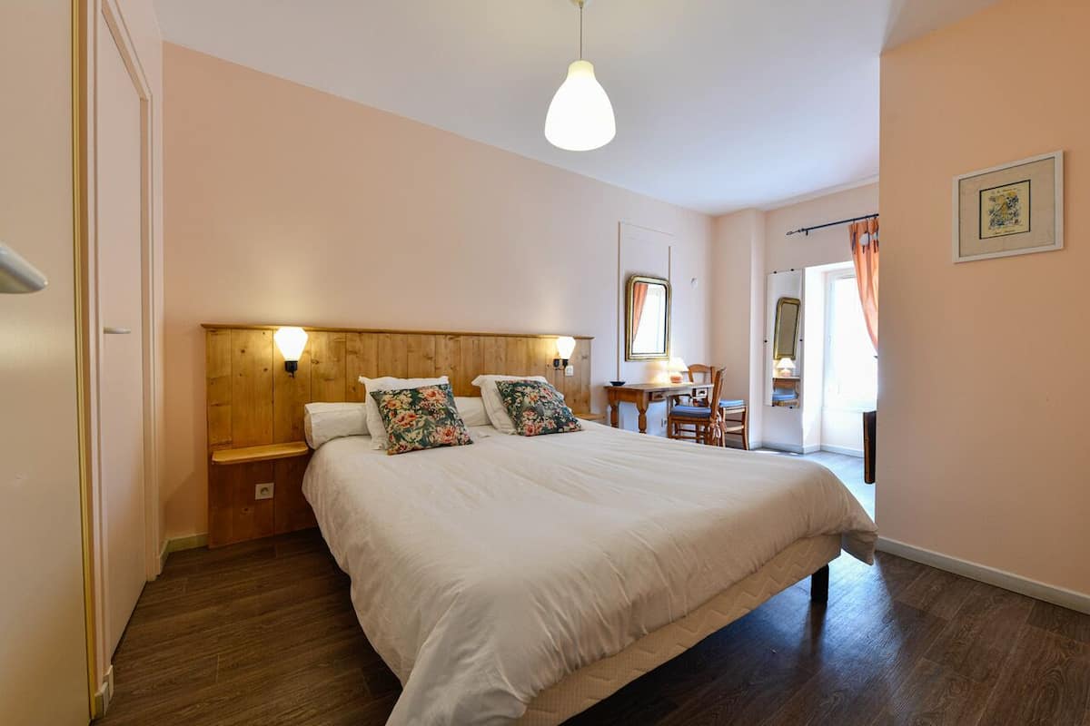 Ein Schlafzimmer mit einem hölzernen Kopfteil, einem großen Bett mit weißer Bettwäsche, zwei Nachttischlampen, einem kleinen Tisch mit Stuhl und einem Fenster mit Vorhängen.
