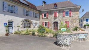 Un edificio de piedra con varias ventanas y contraventanas, un área pavimentada al frente y un pequeño jardín.
