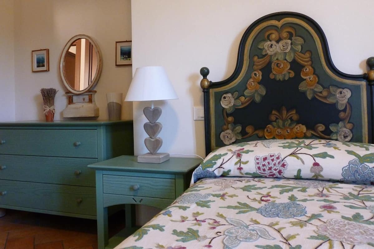 Un dormitorio con una cama de estampado floral, un aparador con cajones, una mesita de noche con lámpara y un espejo en la pared.