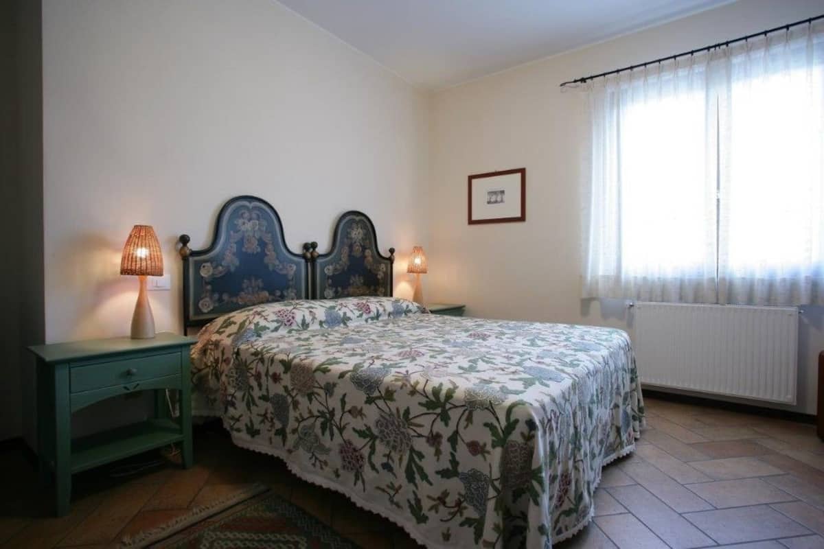 Un dormitorio con una cama, dos lámparas de noche, un radiador y una ventana con cortinas.