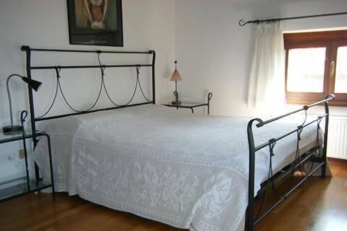 Un dormitorio con estructura de cama metálica, cubrecama blanco, mesita de noche con lámpara y ventana con cortinas.