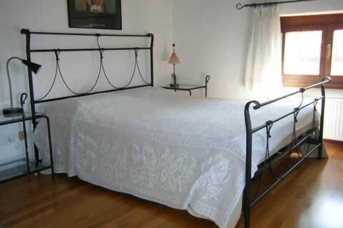 Un dormitorio con estructura de cama metálica, cubrecama blanco, mesita de noche, lámpara y ventana con cortinas.