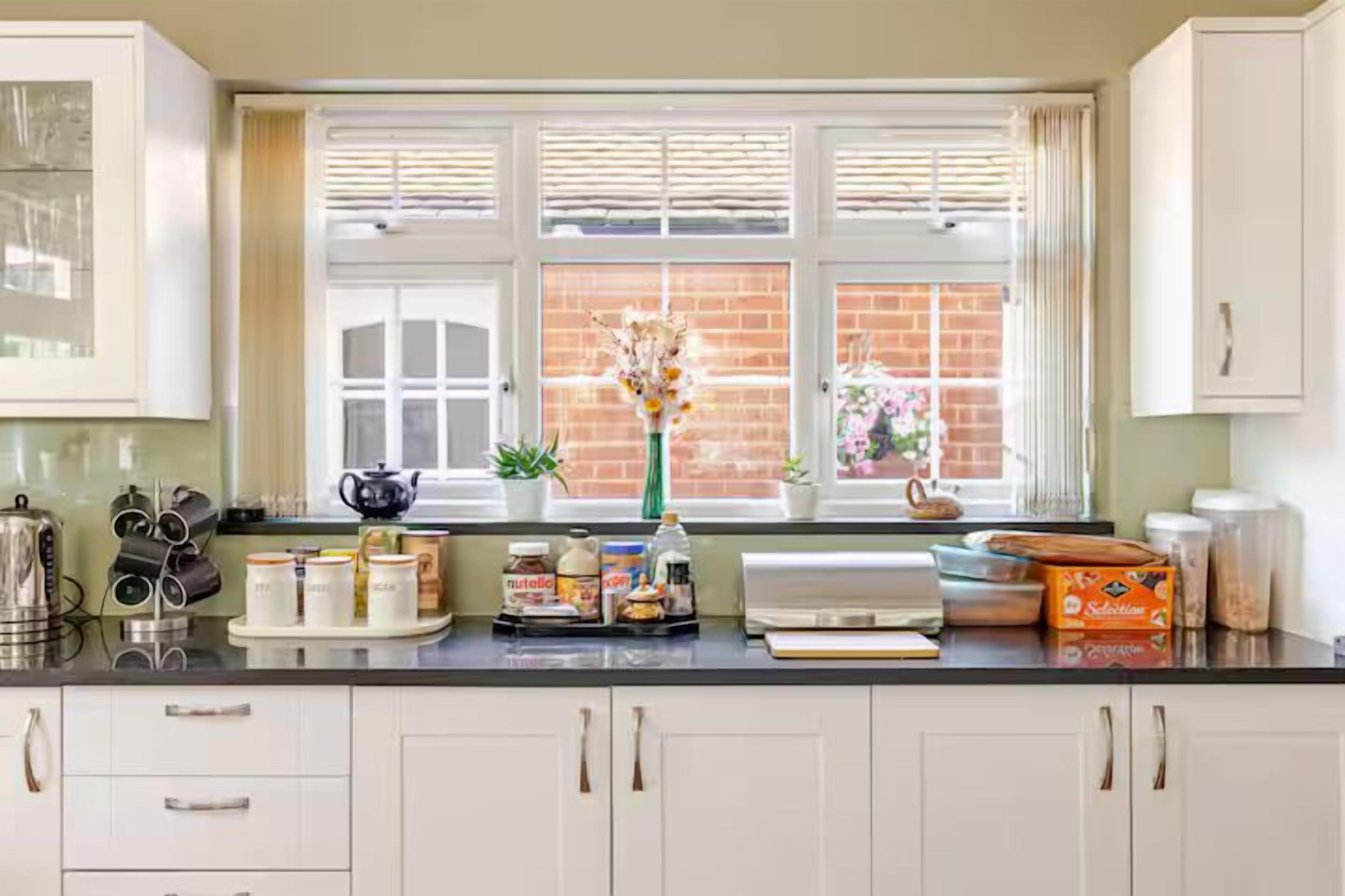 A kitchen with white cabinets, a window with blinds, a vase with flowers, and various kitchen items on the counter.