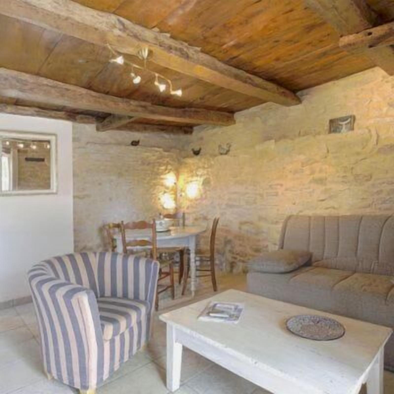 A rustic room with exposed wooden beams, a stone wall, a striped armchair, a sofa, and a small table with a book.