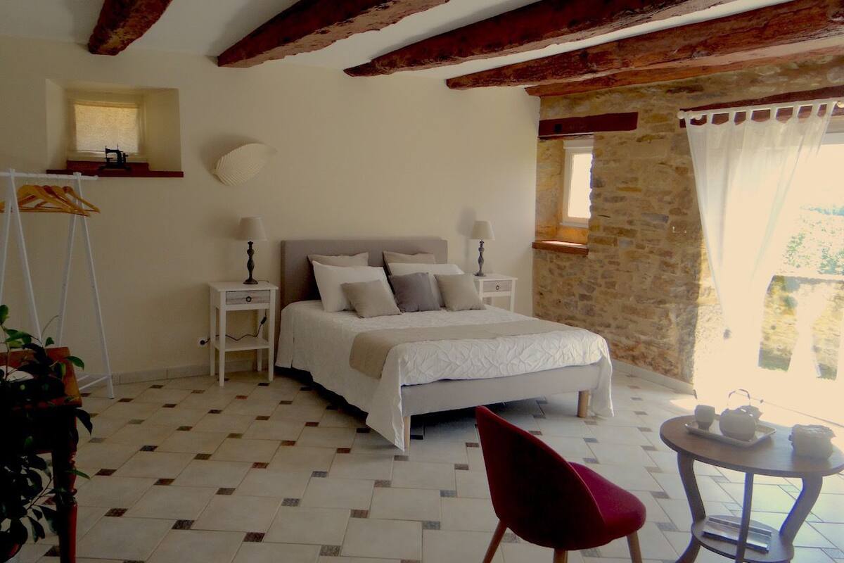 Une chambre à coucher comprenant un lit, des tables de chevet, une chaise, une petite table avec un plateau, et une fenêtre avec des rideaux.