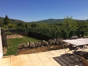 Un patio al aire libre con pared de piedra, una mesa con sillas y vistas a viñedos y colinas.