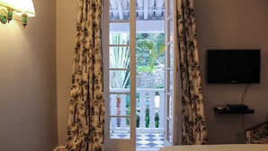 Habitación de hotel con una cama, televisión instalada en la pared y balcón con suelo a cuadros y plantas en macetas.