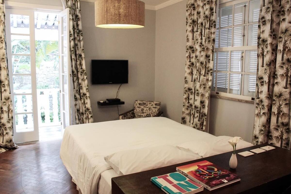 Un dormitorio con cama, televisor montado en la pared, mesita de noche con un libro y un florero, y ventana con cortinas.