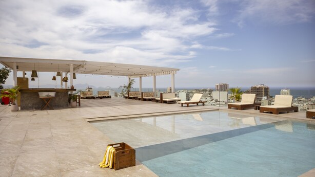 Una piscina en la azotea con sillas de descanso y una pérgola, con vistas a un paisaje urbano y al mar.
