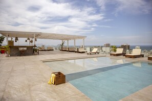 Una piscina en la azotea con sillas de descanso y una pérgola, con vistas a un paisaje urbano y al mar.