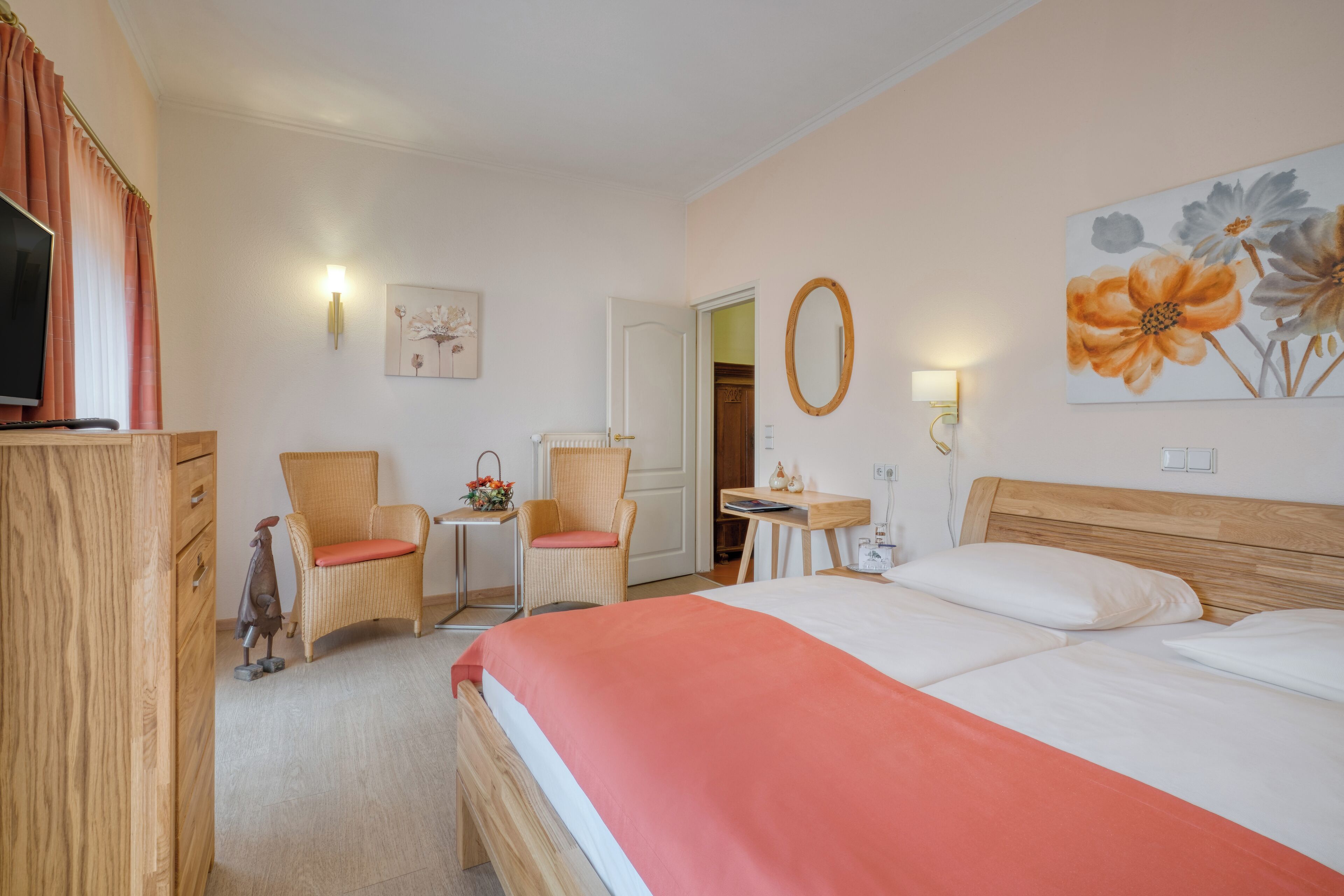 Habitación de hotel con cama, dos sillas, una mesita, cómoda, televisor y obras de arte en las paredes.