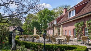 Een terras met tafels en stoelen, omgeven door verzorgde hagen en bomen.