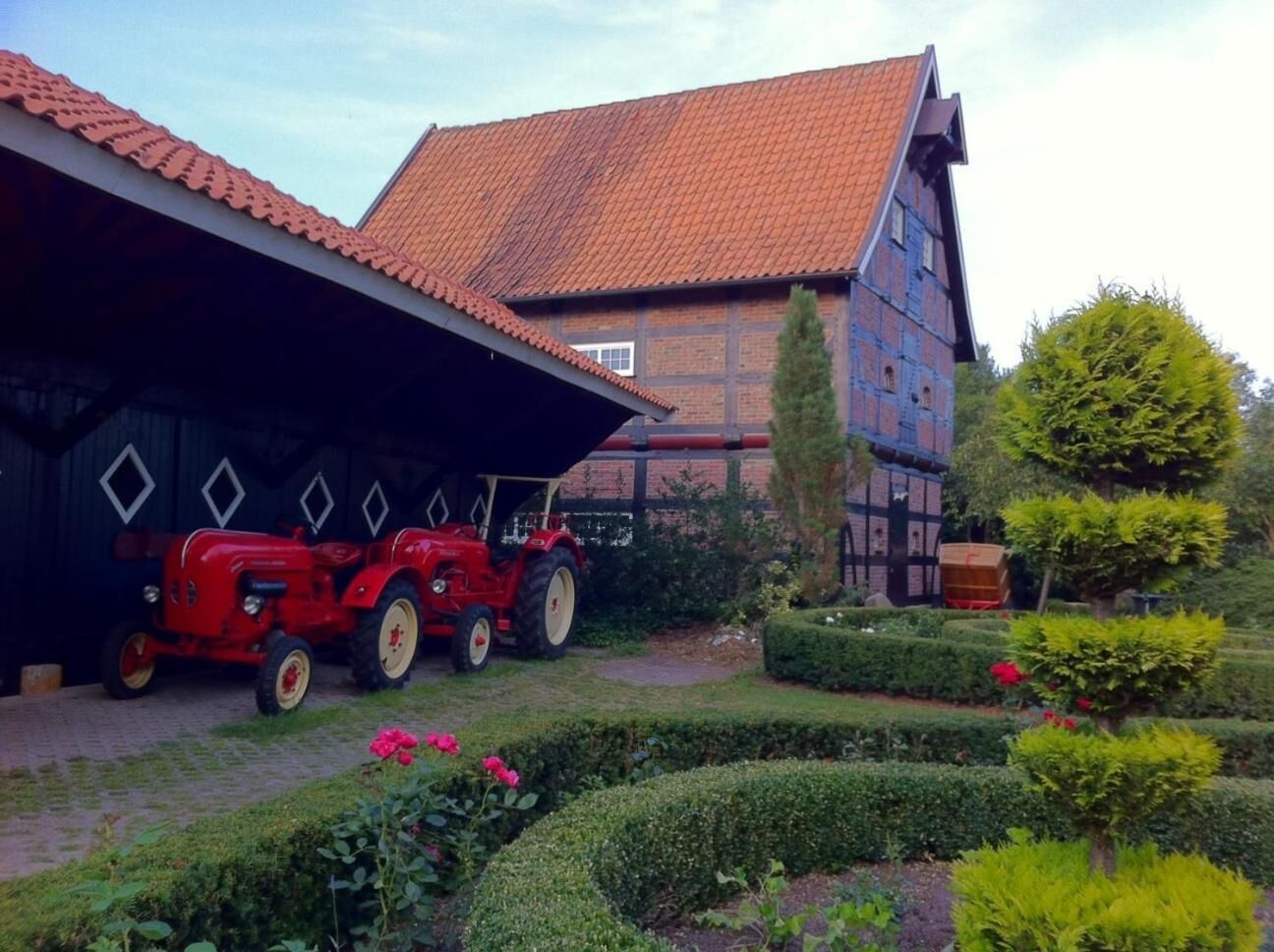 A red tractor parked in front of a barn with a red-tiled roof, surrounded by well-maintained hedges and flowers.