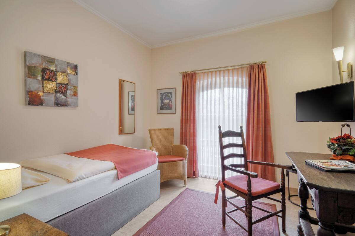 Habitación de hotel con cama, dos sillas, escritorio con lámpara, televisión y ventana con cortinas.