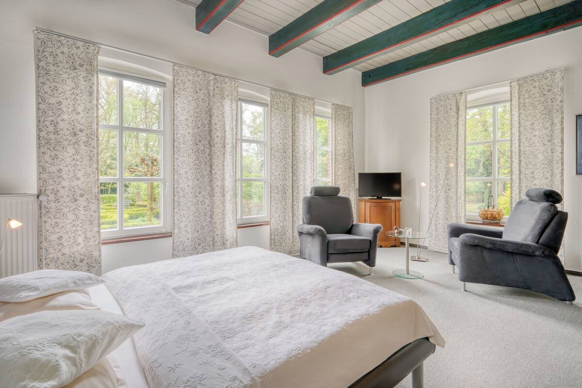 Un dormitorio con una cama grande, dos sillones, un televisor y ventanas amplias con cortinas.