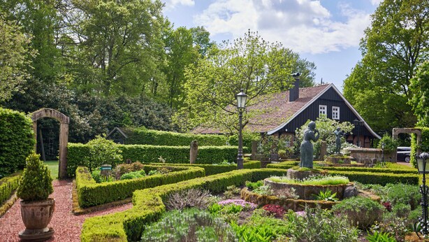 Een verzorgde tuin met een traditioneel huis, strakke hagen en een standbeeld in het midden.