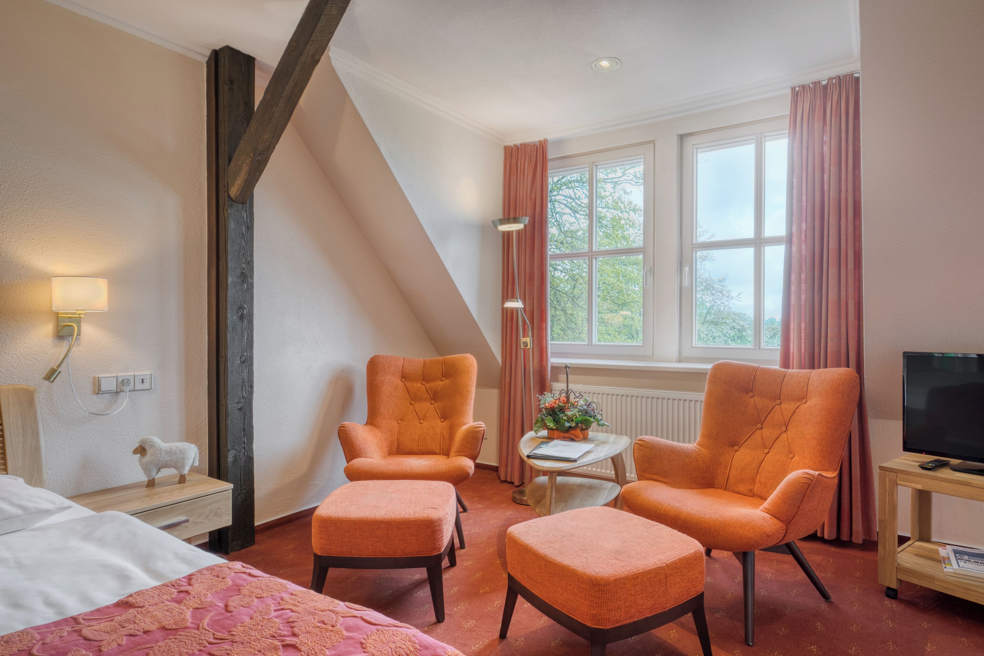 A hotel room with two orange armchairs, a small table with a plant, a bed with a pink blanket, and a TV on a wooden stand.