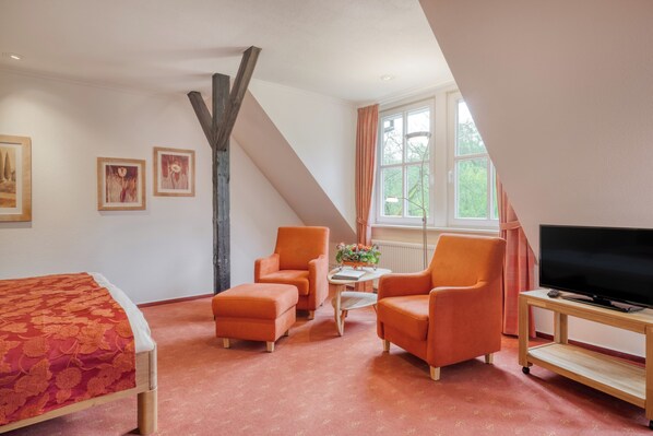 A room with a bed, two armchairs, a small table, a TV, and a window with curtains.