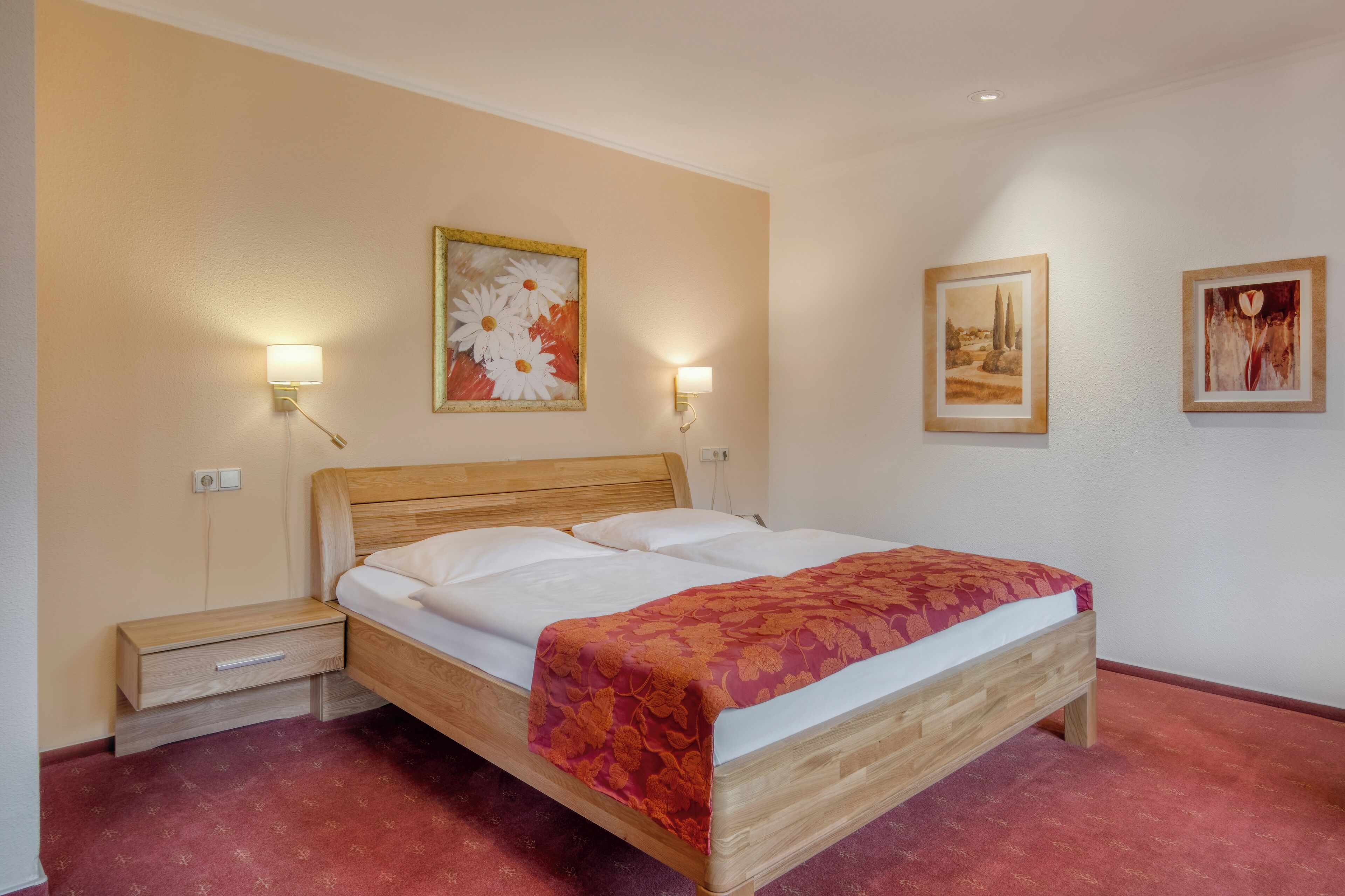 A neatly made bed with a wooden headboard, a nightstand with a drawer, and two wall-mounted lamps.
