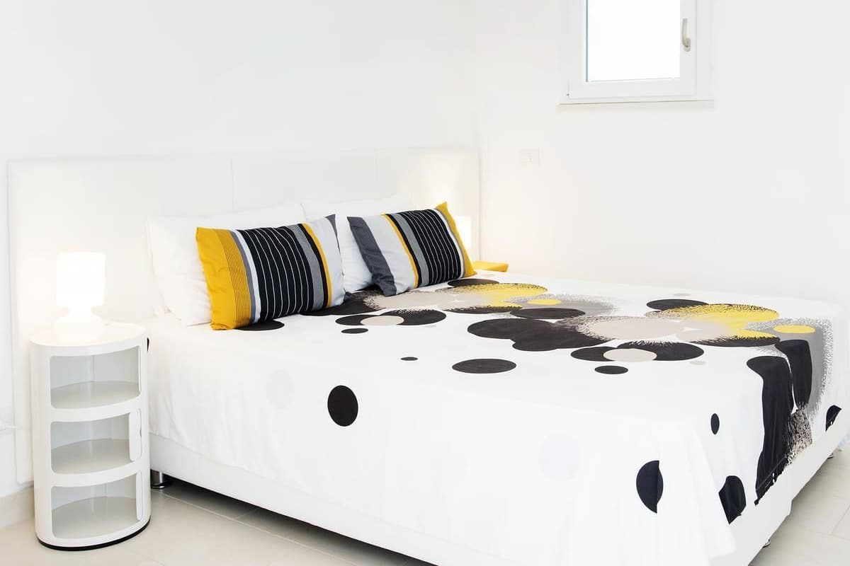 A neatly made bed with a white headboard, a bedside table with a lamp, and a wall-mounted mirror.