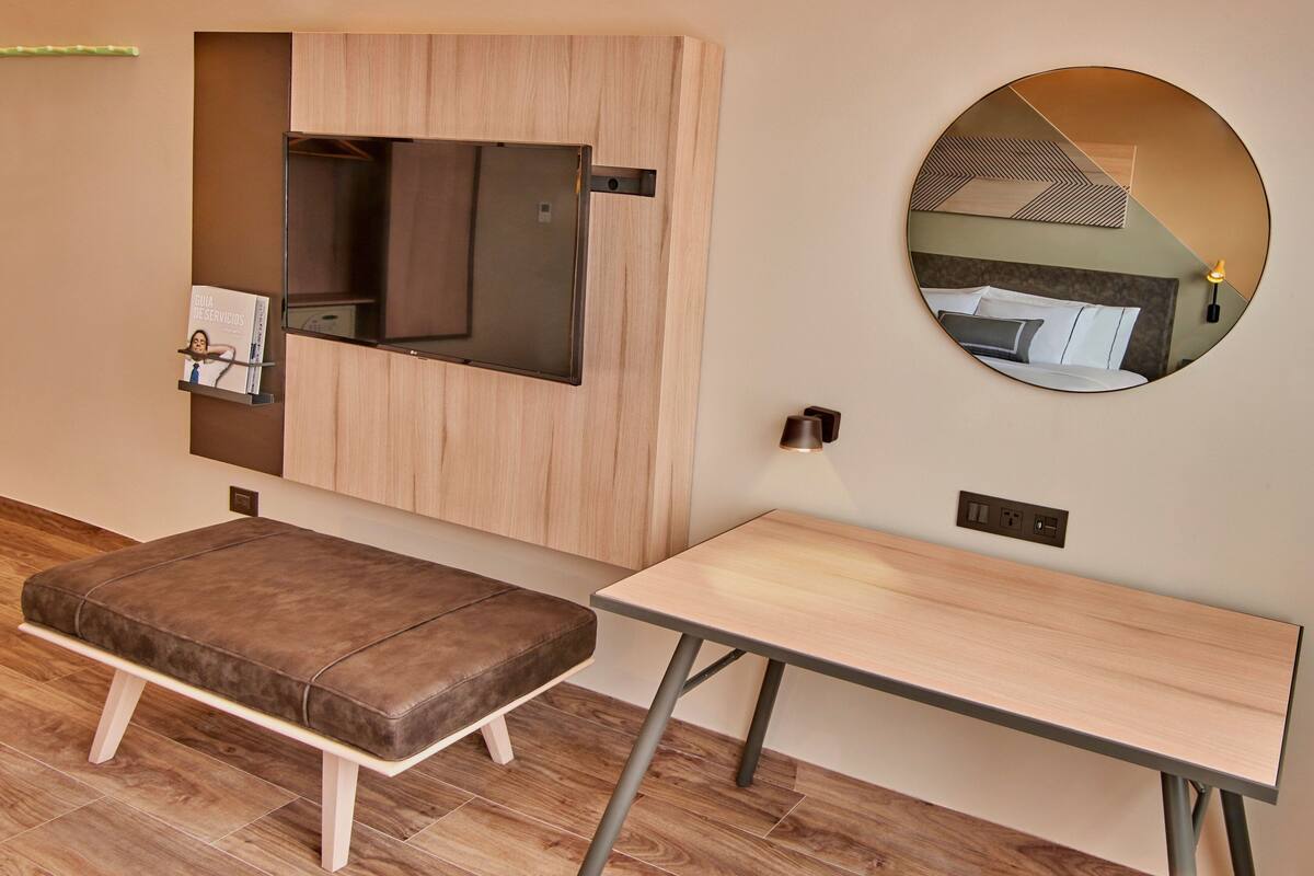 Una habitación moderna con un panel de madera en la pared, televisor montado, un espejo redondo, una pequeña mesa y un banco.