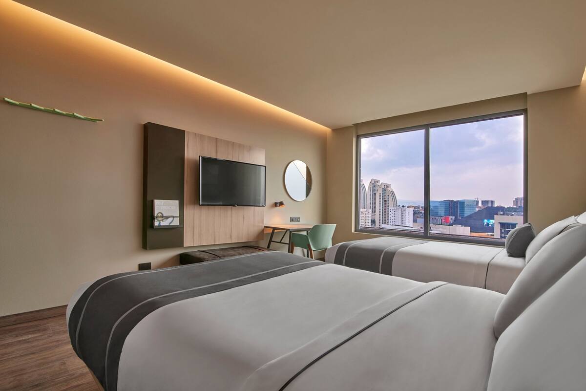 Habitación de hotel moderna con una cama grande, un escritorio, una silla, un televisor y un amplio ventanal con vista a la ciudad.