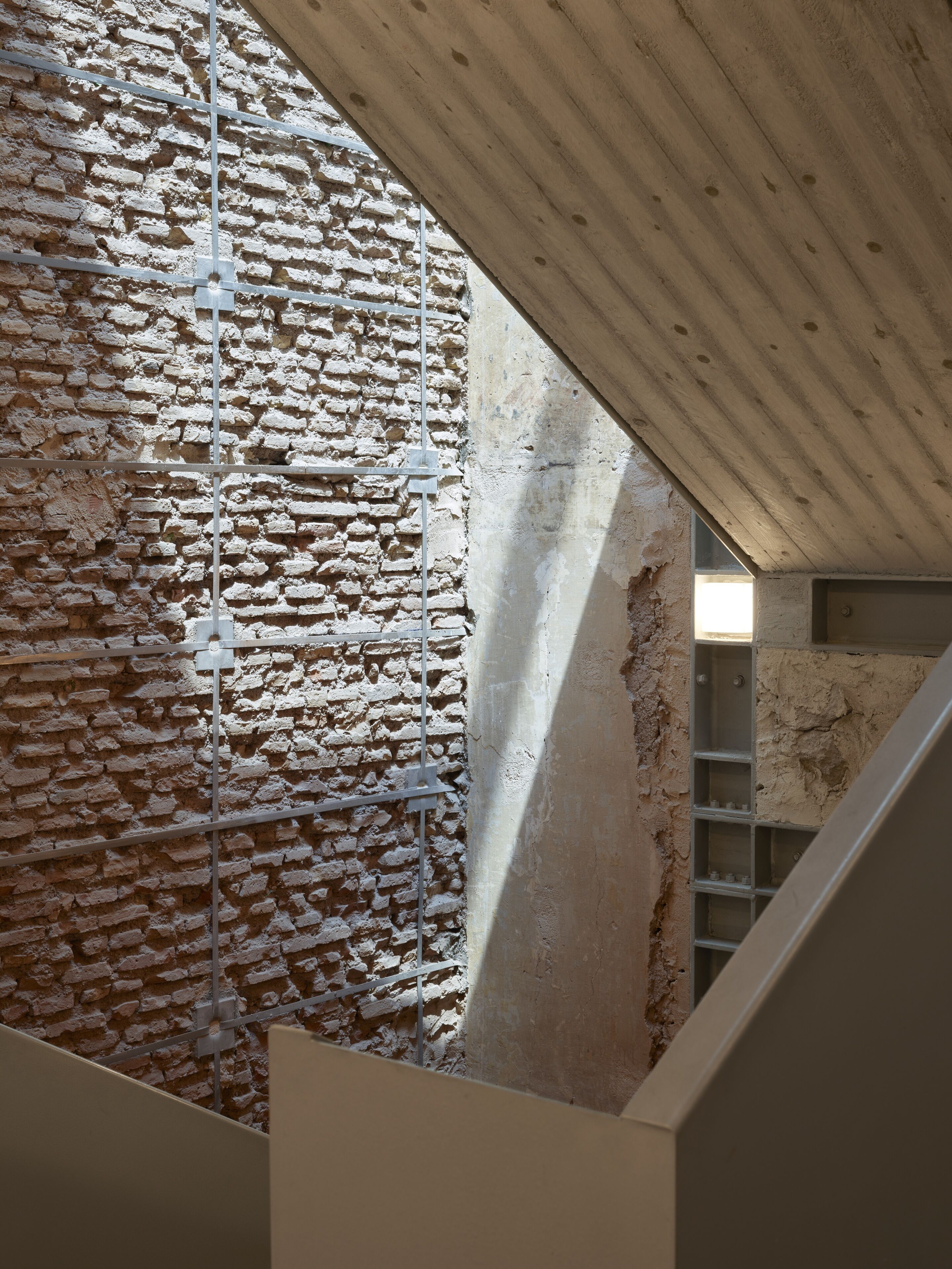A staircase with a brick wall and metal beams, leading to a room with a concrete ceiling and a window.