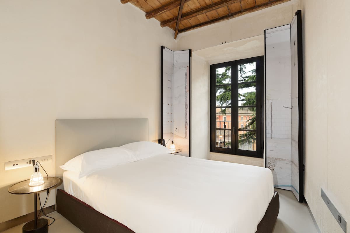 A bedroom with a bed, a bedside table, a lamp, and a window with a view of buildings outside.