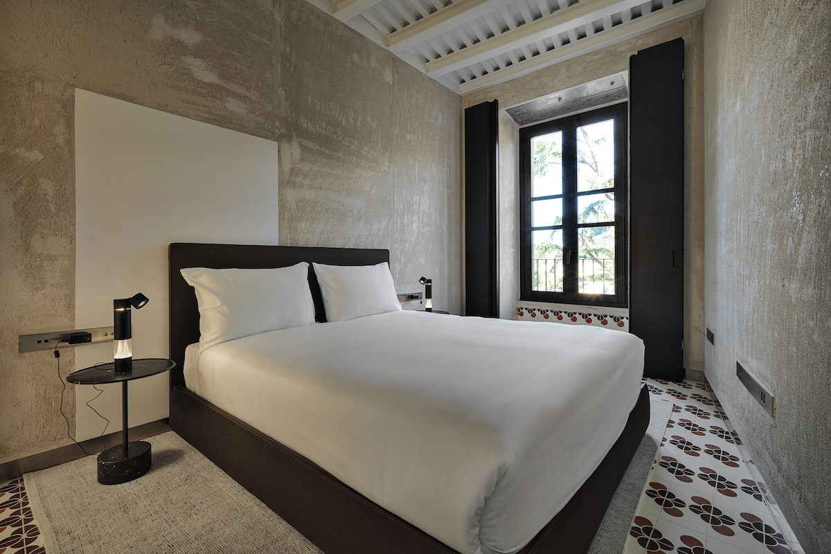 A bedroom with a large bed, a nightstand, a window with curtains, and a patterned rug.