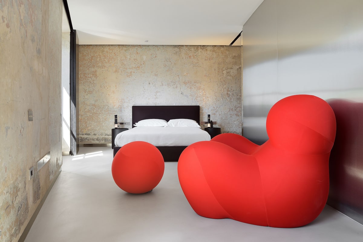 A modern bedroom with a large bed, a concrete wall, and two red bean bags.