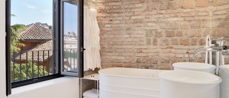 A bathroom with a large bathtub, a window with a view of buildings, and a brick wall.