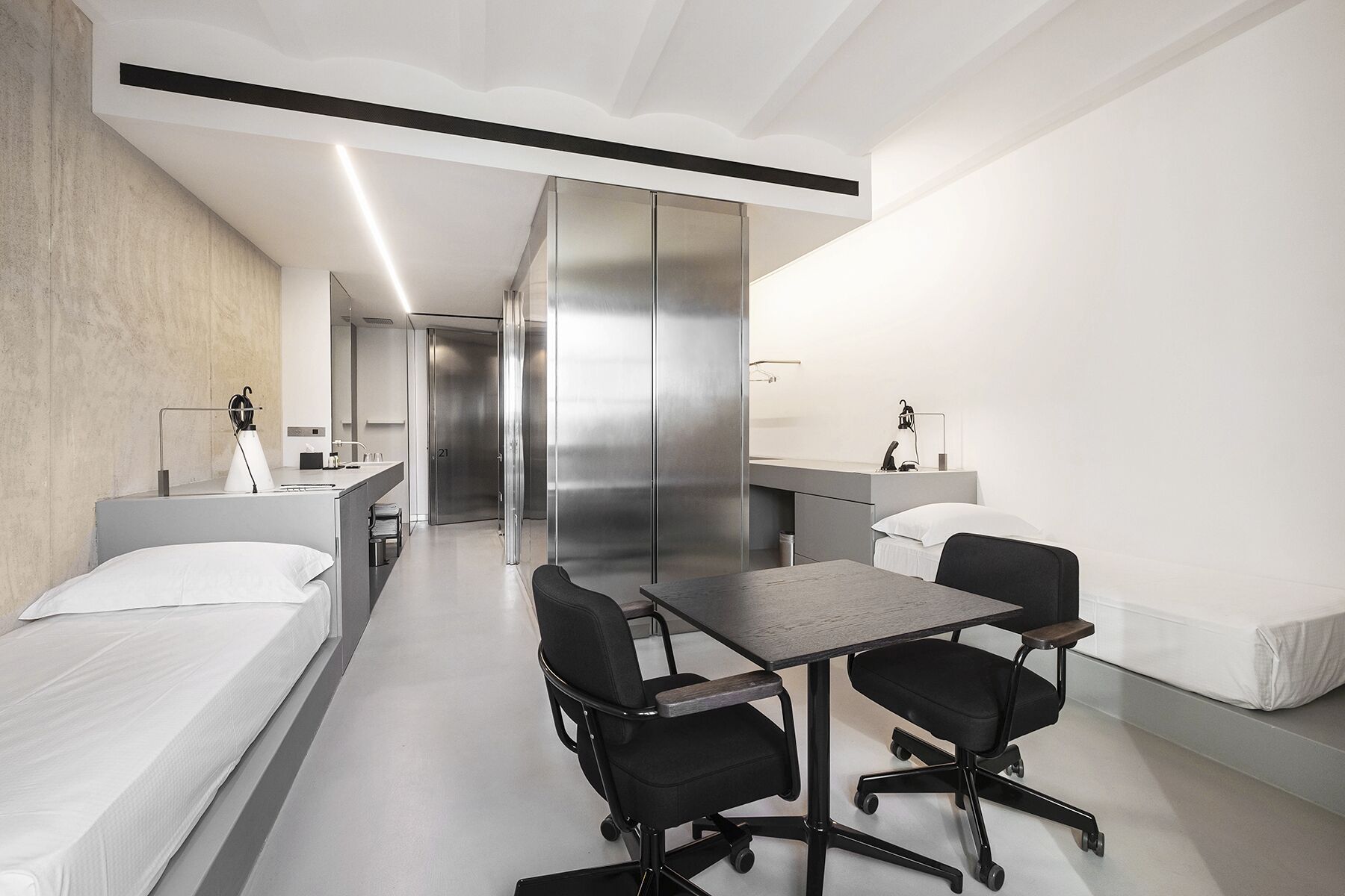 Une chambre d’hôtel moderne équipée d’un lit, d’un bureau, de deux chaises, d’une petite table, d’un miroir et d’une salle de bain avec douche et toilettes.