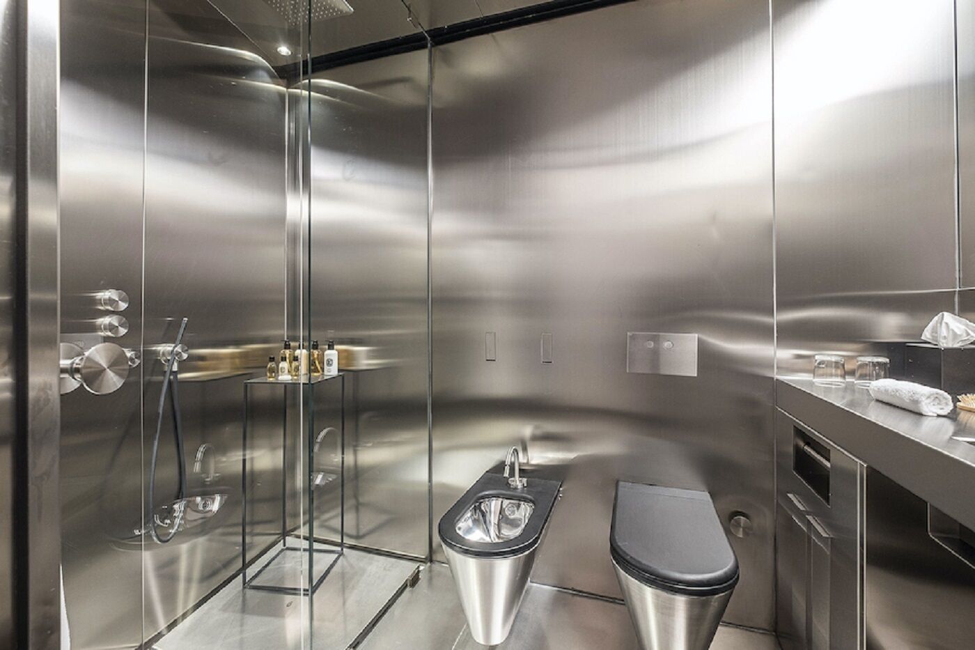 A modern, stainless steel bathroom with a glass shower enclosure, a toilet, and a sink area with toiletries.