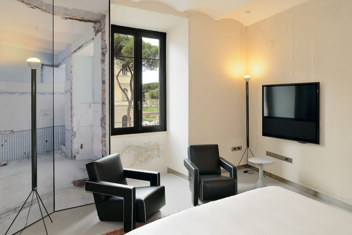 A hotel room with a large bed, two black leather chairs, a TV mounted on the wall, and a window with a view of trees and a building.