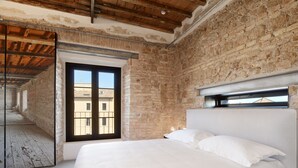 A bedroom with a large bed, a brick wall, and a window overlooking a building. - Rhinoceros (Rome)