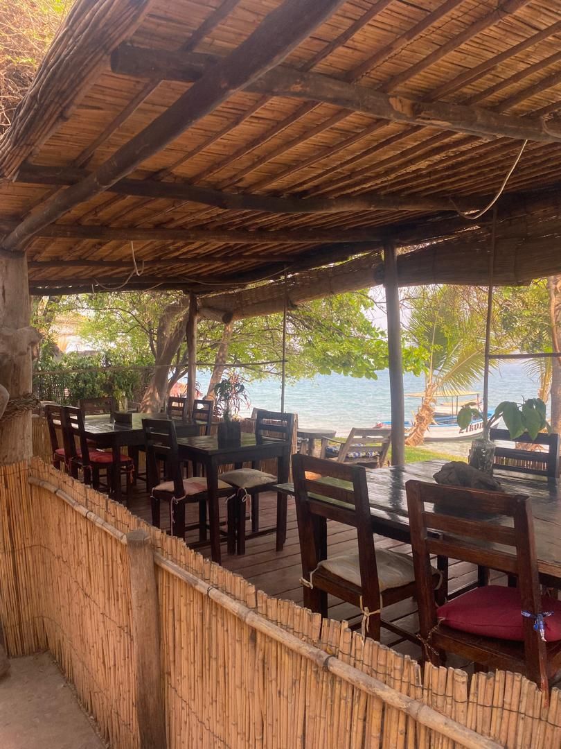 Un área de comedor al aire libre con mesas y sillas de madera, una valla de bambú y vista al mar.