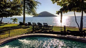 A poolside area with lounge chairs, a pool, and a view of the sea and a distant island. - Mgoza Lodge (Cape Maclear)