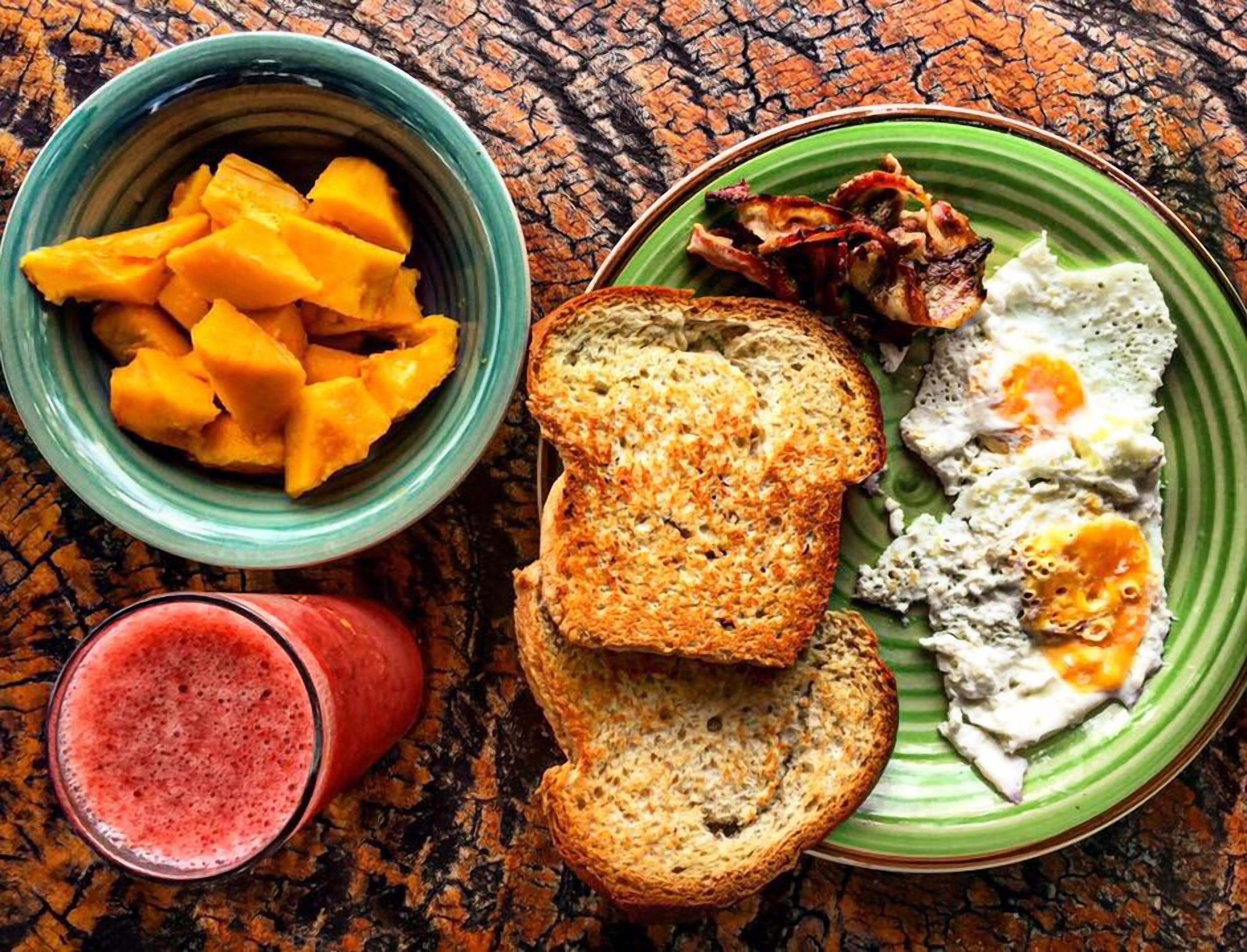 A meal with mangoes, toast, eggs, bacon, and a smoothie on a textured table.